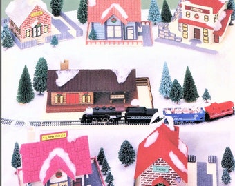 May include: A set of six plastic canvas Christmas village ornaments. The ornaments include a schoolhouse, a general store, a fire station, a train station, a library, and a toy store. Each ornament has a white snowy roof and is decorated with festive details.