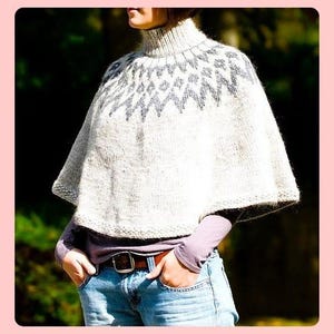 May include: A white knitted poncho with a grey geometric pattern around the neck. The poncho is worn over a purple shirt and blue jeans.