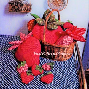 May include: A collection of red and white gingham fabric strawberry pillows. The pillows are arranged in a wicker basket and on a blue and white patterned fabric. The pillows are all different sizes and shapes, and some have green leaves.