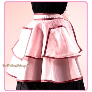 May include: A layered, sheer light pink apron featuring a dark maroon ribbon trim along the edges of the two tiers and the waistband. The apron has two large pockets with maroon trim and a large bow at the back. The text PastPatternPaloozaCo is visible.