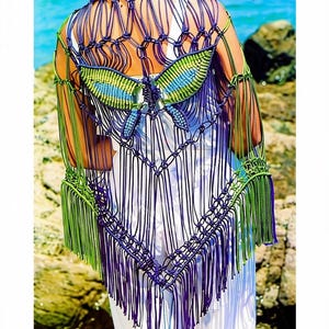 Puede incluir: Un chal de macramé con un diseño de mariposa en morado y verde, patrones de nudos intrincados y flecos largos. La pieza incluye el texto PastPatternPaloozaCo en la esquina superior derecha y se lleva sobre una prenda blanca.