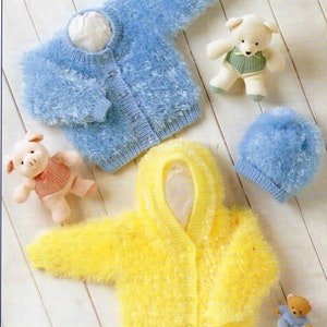 Op de afbeelding: Twee gebreide baby-truien, één blauw en één geel, met een capuchon. De truien zijn gemaakt van een pluche garen en hebben knoopsluitingen. Er zijn ook twee gebreide mutsen en drie knuffels op de afbeelding.