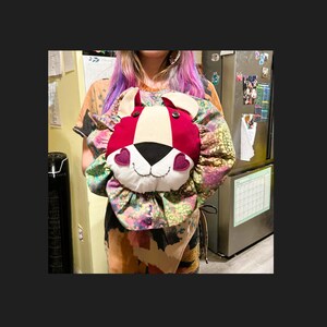 May include: A red and white lion head pillow with black eyes and pink hearts for cheeks. The pillow is surrounded by a colorful floral fabric.