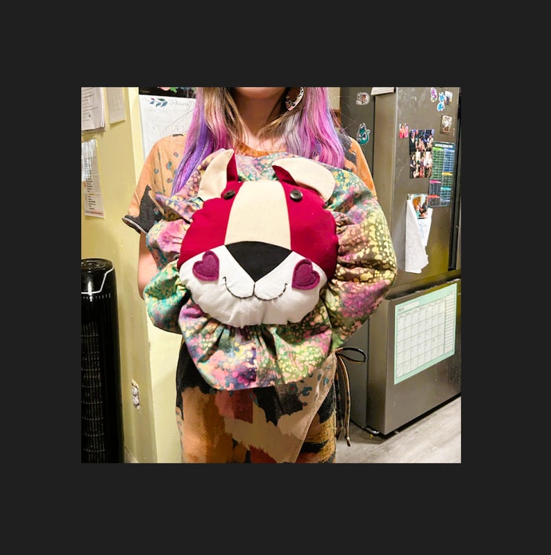 May include: A red and white lion head pillow with black eyes and pink hearts for cheeks. The pillow is surrounded by a colorful floral fabric.