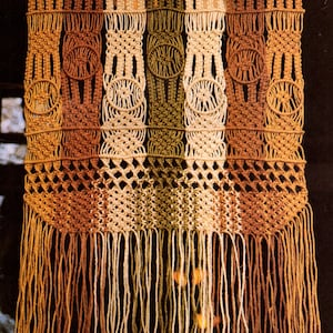 May include: A macrame wall hanging with a geometric pattern in brown, beige, and green yarn. The hanging has a fringe at the bottom.