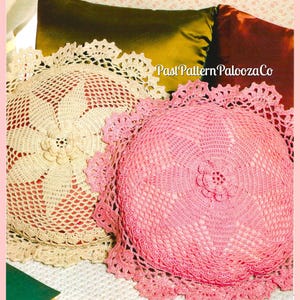 May include: Two crocheted pillow covers, one in beige and one in pink, with a floral design in the center. The pillow covers are on a brown and green surface.