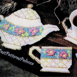 May include: Three white plastic canvas teacup and teapot coasters with a blue, pink, and green floral design. The coasters are on a wooden surface with a book and a cup of coffee.