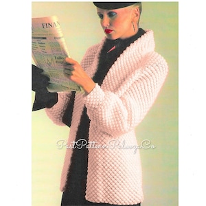 May include: A pink crocheted cardigan with a wide collar. The cardigan is shown being worn by a model holding a newspaper.