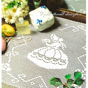 May include: A white crocheted lace doily with a floral design is displayed on a wooden surface. A small white box with blue bow accents sits on the doily, along with a perfume bottle and a bouquet of white flowers. The text "PastPatternPaloozaCo" is visible.
