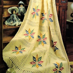 Op de afbeelding: Een gehaakt deken met een franje rand en een herhalend patroon van kleurrijke sterbursts. Het deken is gedrapeerd over een houten trap en een kleine tafel.