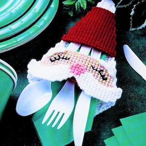 May include: A white crocheted Santa Claus napkin holder with a red hat. The holder is holding a white plastic fork, spoon, and knife. The holder is sitting on a green napkin.