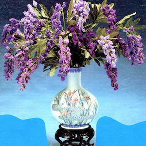 May include: A white ceramic vase with a blue and gold floral design, filled with a bouquet of purple crocheted flowers. The vase is on a wooden stand.