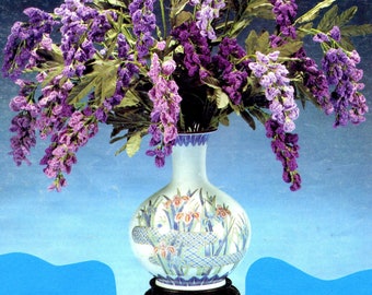 May include: A white ceramic vase with a blue and gold floral design, filled with a bouquet of purple crocheted flowers. The vase is on a wooden stand.