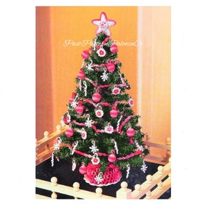 Puede incluir: Un árbol de Navidad de ganchillo decorado con adornos rosas y blancos, una estrella rosa y blanca en la parte superior y una base de ganchillo rosa y blanca. El árbol está sobre una valla blanca de madera.