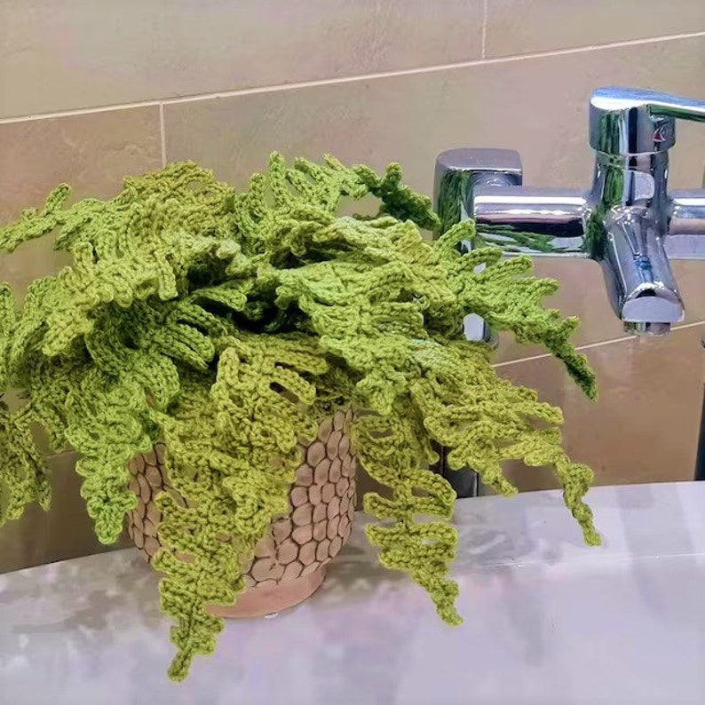 K&ouml;nnte beinhalten: Eine geh&auml;kelte gr&uuml;ne Farnpflanze in einem braunen und beigefarbenen Topf. Die Pflanze steht auf einer wei&szlig;en Arbeitsplatte neben einem verchromten Wasserhahn.