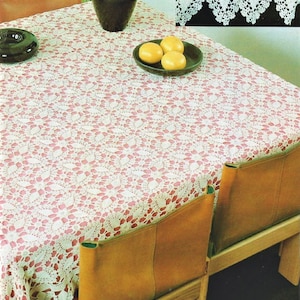May include: A white crocheted tablecloth with a delicate floral pattern, laid over a wooden table with two brown leather chairs. The tablecloth has a pink undertone and a scalloped edge. Three yellow lemons are on a brown plate on the table.