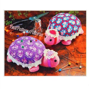 May include: Three handmade turtle pin cushions. The turtle shells are covered in floral fabric, with white lace trim. The turtles are pink with black eyes. Pins are inserted into the purple turtle. The image includes the text "Past Patterns Palooza".