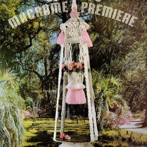 May include: A white macrame hanging planter with pink tassels and a pink bird of paradise flower arrangement. The planter is hanging from a white macrame rope and is set against a backdrop of a lush green garden.