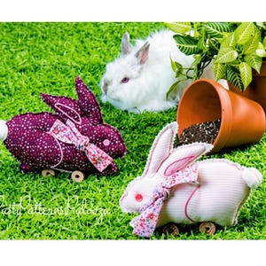 Könnte beinhalten: Zwei Stoffhasen-Spielzeuge auf Rädern, eines in Dunkelviolett mit weißem Blumenmuster und eines in Hellrosa mit dezenten Streifen, beide mit einer Blumen-Schleife, Knopfaugen und einem weißen Schwanz, mit dem Text PastPatternsPalooza.