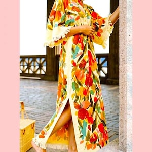 May include: A floor-length kaftan dress featuring a vibrant floral pattern in shades of red, orange, yellow, and teal on a cream background. The garment has wide sleeves, a side slit, and decorative fringe trim along the hem and sleeve edges.