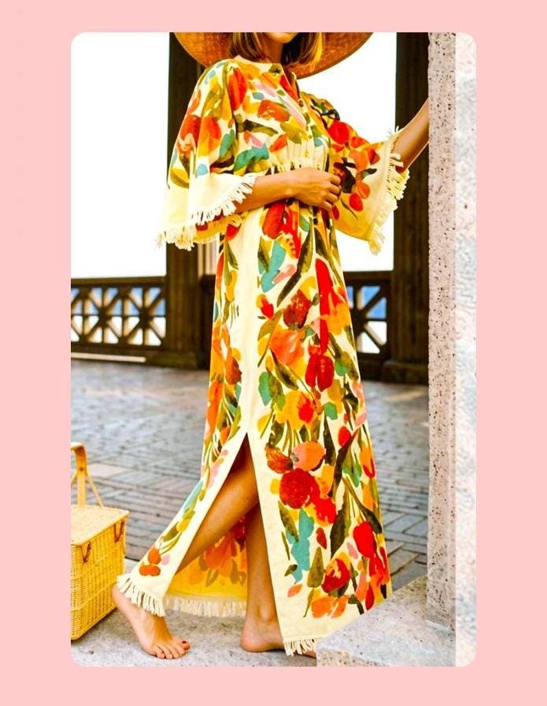 May include: A floor-length kaftan dress featuring a vibrant floral pattern in shades of red, orange, yellow, and teal on a cream background. The garment has wide sleeves, a side slit, and decorative fringe trim along the hem and sleeve edges.