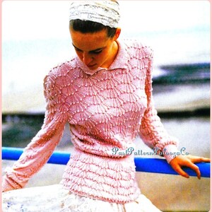 以下が含まれることがあります： 淡いピンク色のニットセーター。扇のような模様が施されています。長袖のトップスは襟付きで、白いスカートと合わせています。画像はピンク色の枠で囲まれています。