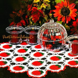 May include: White crocheted table runner with red and green apples. The runner is on a dark wood table with three glass candle holders and a glass pumpkin-shaped container.