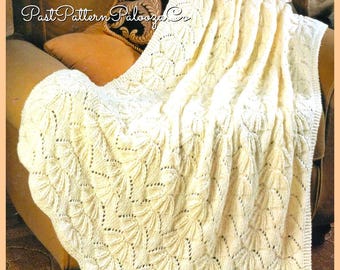 May include: Cream-coloured knitted blanket with a detailed leaf pattern draped over a chair. The blanket has a scalloped edge and is made of a soft, textured yarn. The chair has a brown cushion and a decorative pillow.