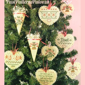 K&ouml;nnte beinhalten: Eine Sammlung cremefarbener Kreuzstich-Ornamente, die an einem gr&uuml;nen Baum h&auml;ngen. Die Ornamente umfassen Herzformen und Kegel mit gestickten Motiven von Glocken, Stechpalmen und dem Text We wish you a Merry Christmas and a Happy New Year.