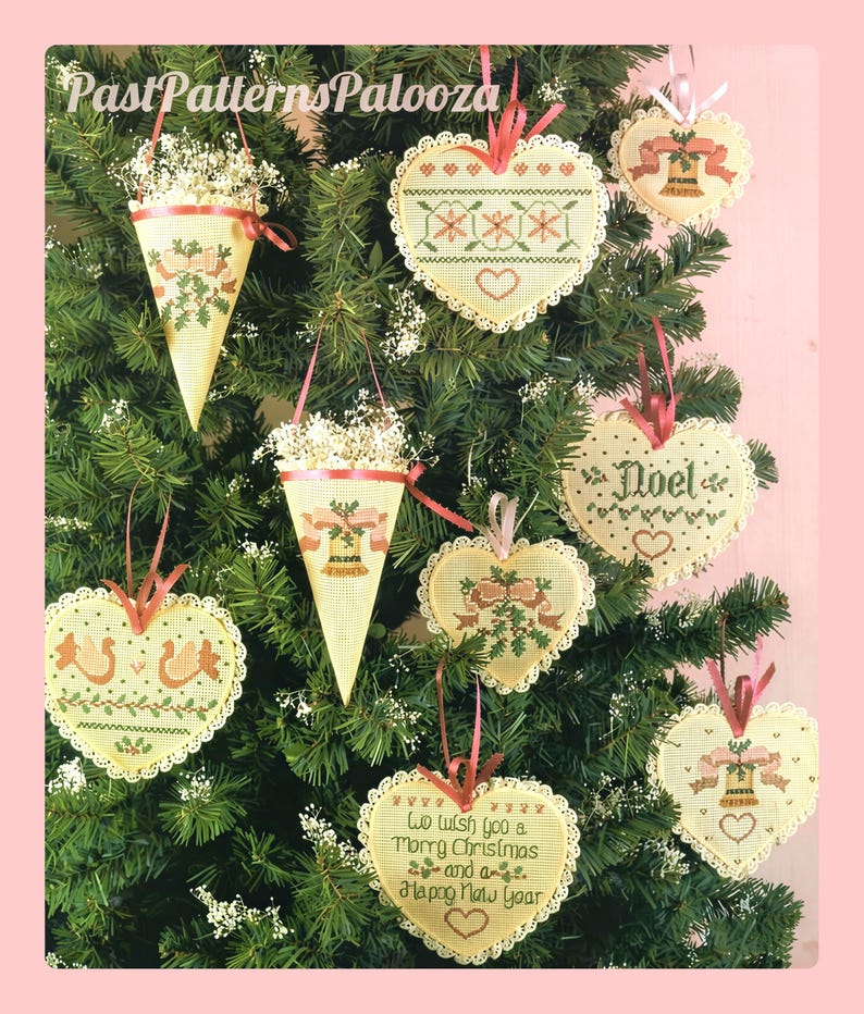K&ouml;nnte beinhalten: Eine Sammlung cremefarbener Kreuzstich-Ornamente, die an einem gr&uuml;nen Baum h&auml;ngen. Die Ornamente umfassen Herzformen und Kegel mit gestickten Motiven von Glocken, Stechpalmen und dem Text We wish you a Merry Christmas and a Happy New Year.