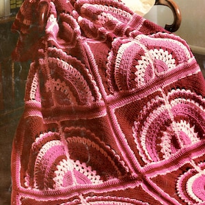May include: A crocheted blanket with a pink and burgundy color scheme. The blanket is made up of squares with a fan-shaped design in the center.
