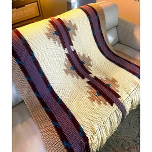Peut inclure: Couverture en crochet cr&egrave;me avec un motif g&eacute;om&eacute;trique dans les tons de violet, bordeaux et beige. La couverture a une bordure &agrave; franges et est drap&eacute;e sur un canap&eacute;. Le motif pr&eacute;sente des formes de losanges r&eacute;p&eacute;t&eacute;es.