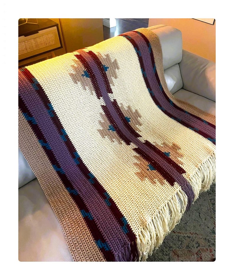 Peut inclure: Couverture en crochet cr&egrave;me avec un motif g&eacute;om&eacute;trique dans les tons de violet, bordeaux et beige. La couverture a une bordure &agrave; franges et est drap&eacute;e sur un canap&eacute;. Le motif pr&eacute;sente des formes de losanges r&eacute;p&eacute;t&eacute;es.