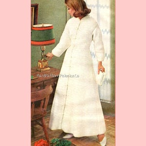 May include: A woman in a long, white, button-down coat dress. The dress has a textured pattern and is paired with white shoes and a small white clutch. A lamp with a green shade and red trim sits on a wooden table. The text "PastPatternPaloozaCo" is visible.