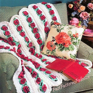 May include: A white crocheted blanket with red roses and green leaves draped over a green armchair. A floral pillow with red roses and two red books are also on the chair. The blanket has a scalloped edge.
