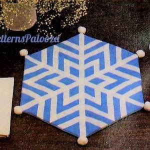 May include: A blue and white snowflake-shaped placemat with white pom-poms around the edges. The placemat is on a dark brown wooden table with a white napkin, fork, knife, and spoon.