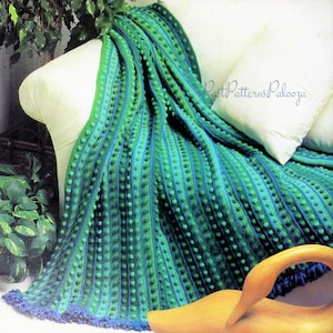 May include: A handmade, textured blanket in shades of green and blue, draped over a white sofa. The blanket features vertical stripes with small, raised bobbles, and a scalloped edge. The overall design is reminiscent of a cozy, handcrafted item.