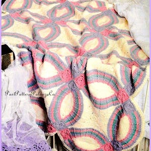 Puede incluir: Una manta de ganchillo hecha a mano con un diseño floral. La manta presenta un fondo crema con grandes formas de flores en rosa, gris y turquesa. La manta está sobre un marco de cama.