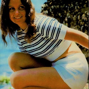 Puede incluir: Una mujer con un top de punto blanco y azul a rayas y una falda de punto blanca. Está sentada en una superficie rocosa con un cielo azul de fondo.