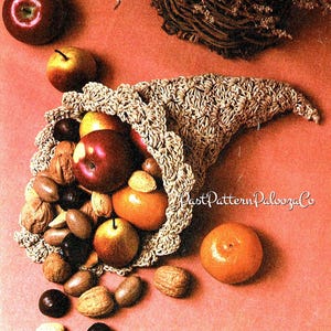 May include: A beige crocheted cornucopia filled with apples, oranges, and nuts. The horn of plenty sits on a peach-coloured surface. A dark red apple and a small orange are also visible.