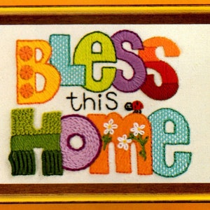 Op de afbeelding: Een kleurrijk geborduurd wandkleed met de woorden "Bless this Home" in een grillig lettertype. De letters zijn geborduurd in verschillende kleuren, waaronder geel, groen, paars, oranje en blauw. Een klein lieveheersbeestje is geborduurd boven het woord "Home" en er zijn witte bloemen geborduurd onder het woord.