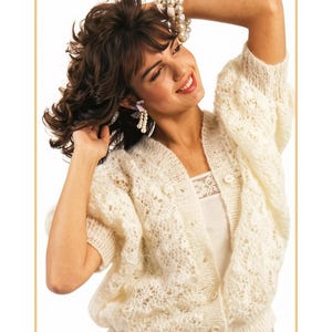 May include: A cream-colored, open-knit cardigan featuring a delicate lace-like pattern, short sleeves, a V-neckline, and a front button closure. The garment is worn over a white camisole with lace trim. The text PastPatternPaloozaCo is visible.