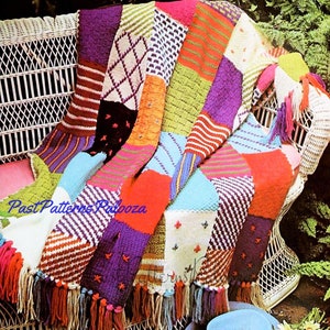 May include: A colorful knitted patchwork throw blanket with fringe. The blanket is made up of squares in various colors, including red, orange, yellow, green, blue, purple, and white. The blanket is draped over a white wicker chair.