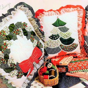May include: Christmas-themed pillows with white backgrounds, adorned with appliqued wreaths, Christmas trees, and bows. The pillows are trimmed with lace and polka dot fabric. A basket of spools and wrapped gifts are also visible.