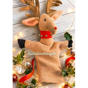 May include: A tan fleece reindeer hand puppet featuring large antlers, a red collar with small gold bells, a red nose, and dark brown cuffs at the ends of the arms. The text "PatiPottersPaloogaCo" is printed in white across the center of the puppet.