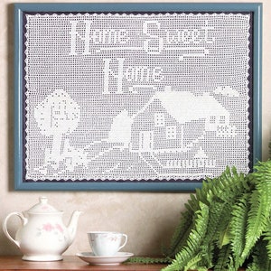 May include: A framed white crocheted piece of art with a blue frame. The crocheted piece depicts a house with a fence and a tree. The frame is hanging on a wall with a white teapot, a teacup and saucer, and a green fern plant on a wooden surface below.