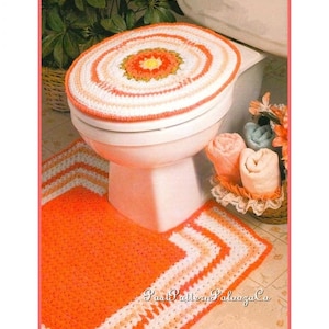 Könnte beinhalten: Ein gehäkeltes WC-Sitzbezug- und Teppichset in Orange-, Weiß- und Rosatönen. Der Sitzbezug hat ein kreisförmiges Design mit einer Blume in der Mitte. Der Teppich hat einen weißen und orangefarbenen Streifenrand.