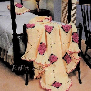 May include: A crocheted throw blanket with a pink and white floral design. The blanket is made with a square pattern and has a ruffled edge. The blanket is draped over a bed with a wooden headboard.