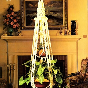 May include: A white macrame plant hanger with a brown pot and green plant hanging from it. The hanger is in front of a fireplace with a painting above it. The title "Soft On Macrame" is at the top of the image.
