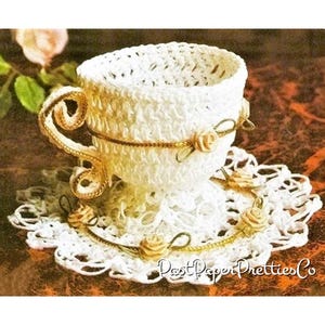 May include: White crocheted teacup and saucer set with gold trim and small white crocheted roses. The set is on a brown background.